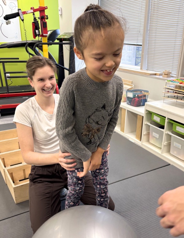 A physiotherapist working with a child on their Osteogenesis Imperfecta. The child is standing up in front of a ball while the physiotherapist sits behind them steadying them by holding their legs. Both are smiling.